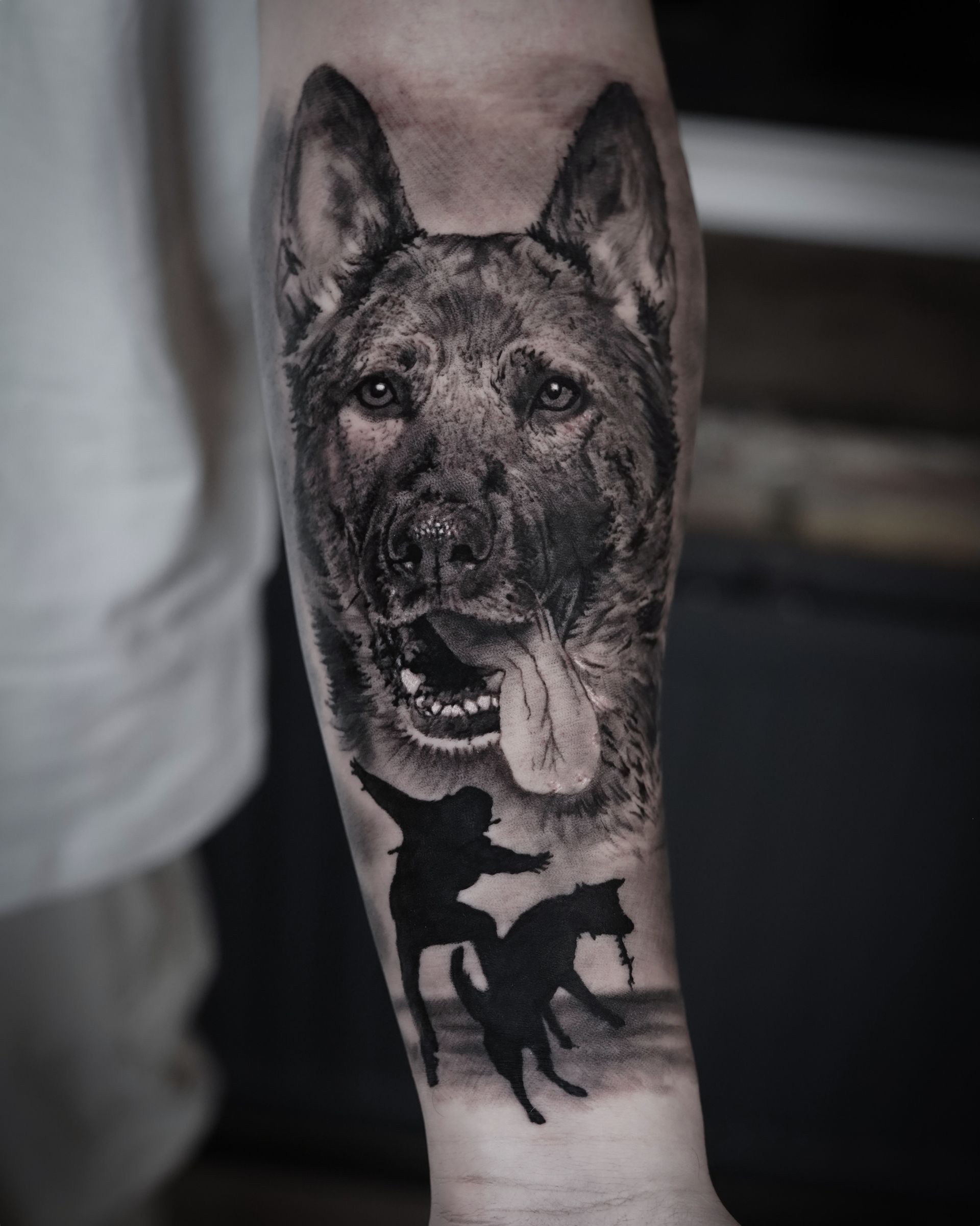 A black and white tattoo of a dog and a man on a forearm.