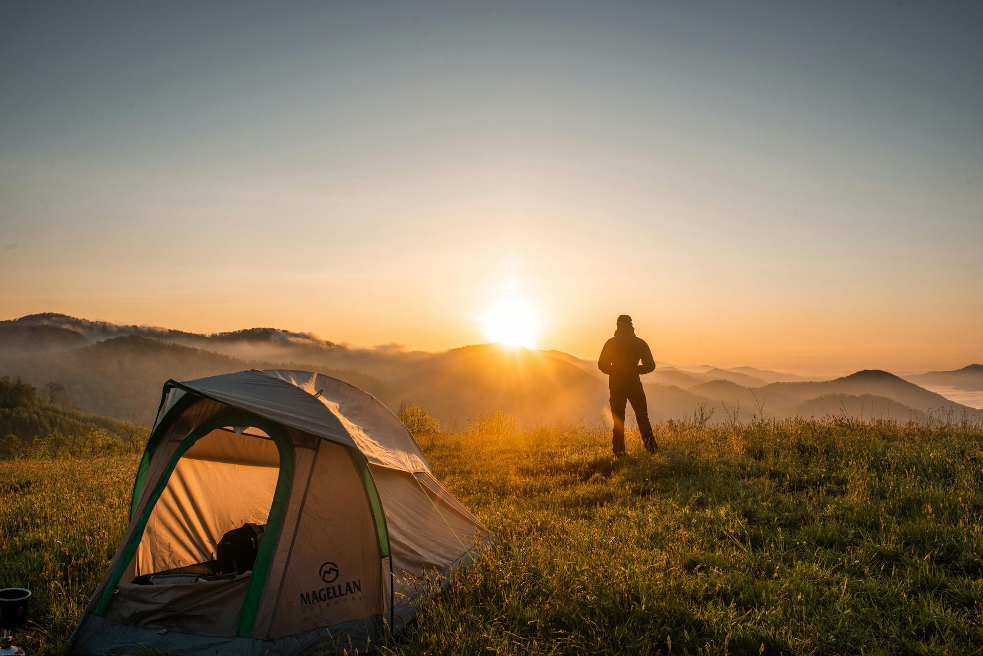Backpacker beside a tent in a grassy field at sunrise, with mountains in the distance.