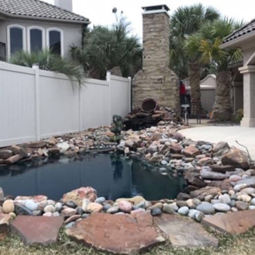 A backyard with a pond surrounded by rocks and a white fence.