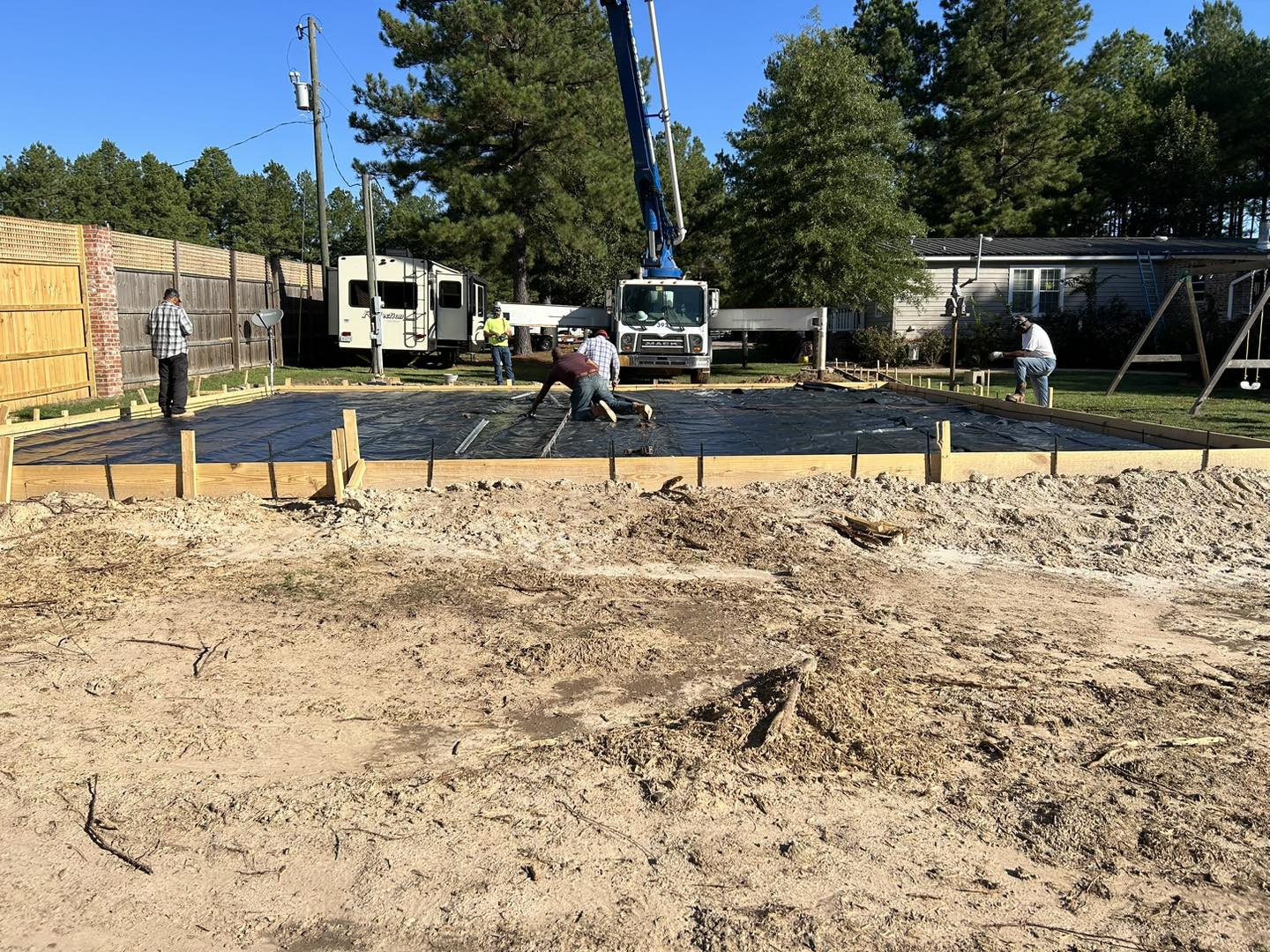 Workers pouring concrete into a wooden frame, setting: outdoors. Concrete truck, dirt, surrounding buildings.