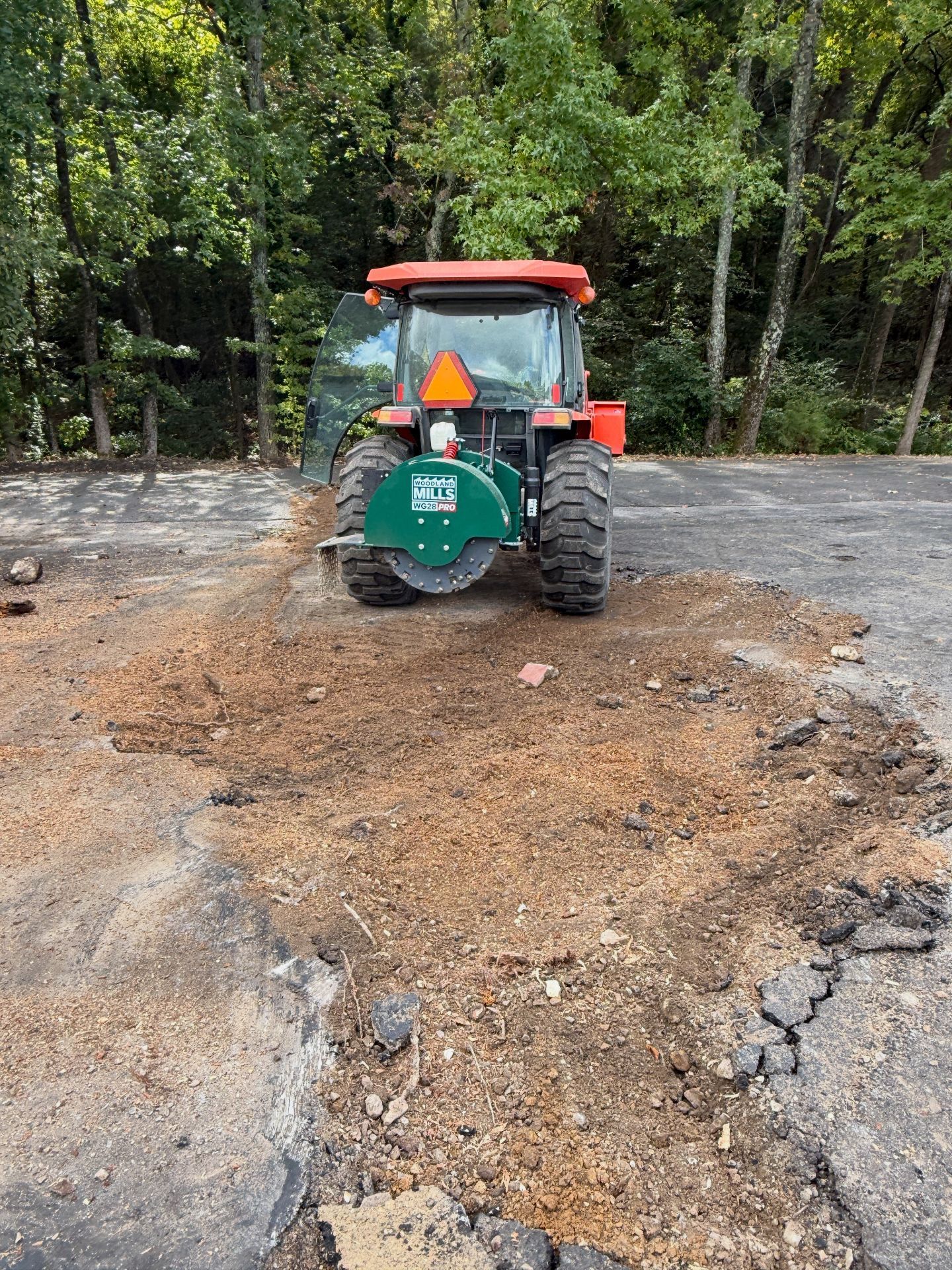 Orange tractor with stump grinder removing asphalt, trees in background.