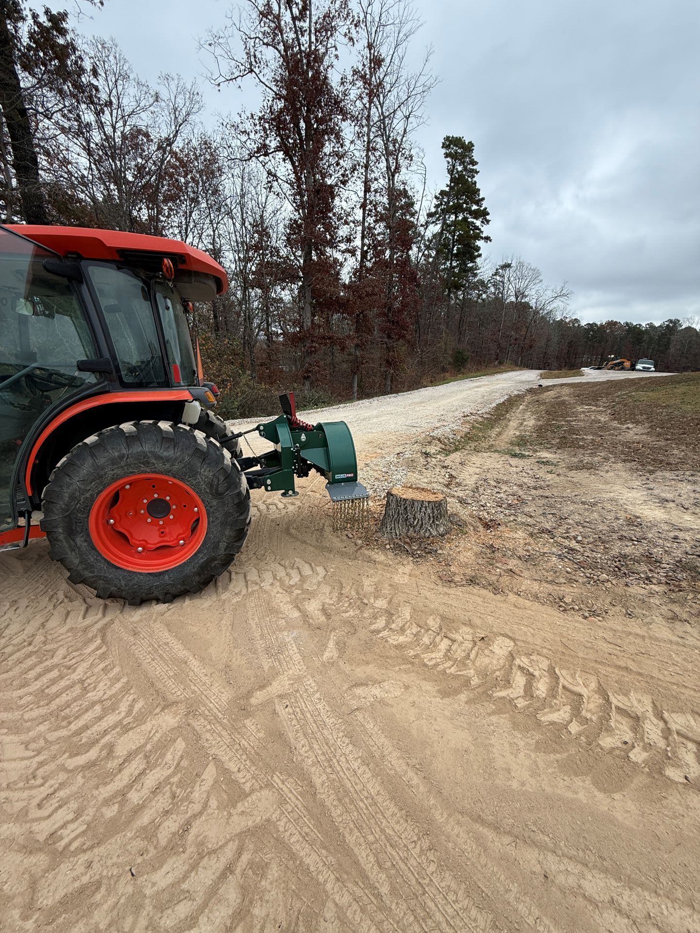 Orange tractor with brush cutter grading a gravel road in a wooded area.