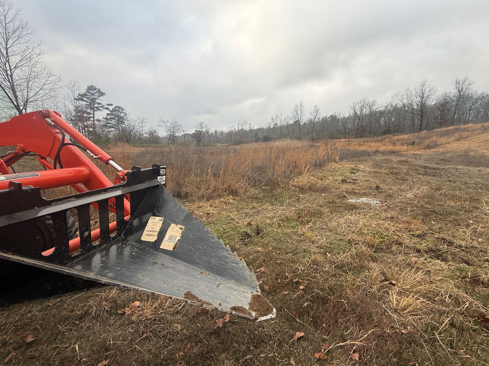 Orange tractor bucket in a field of brown grass, under a cloudy sky.