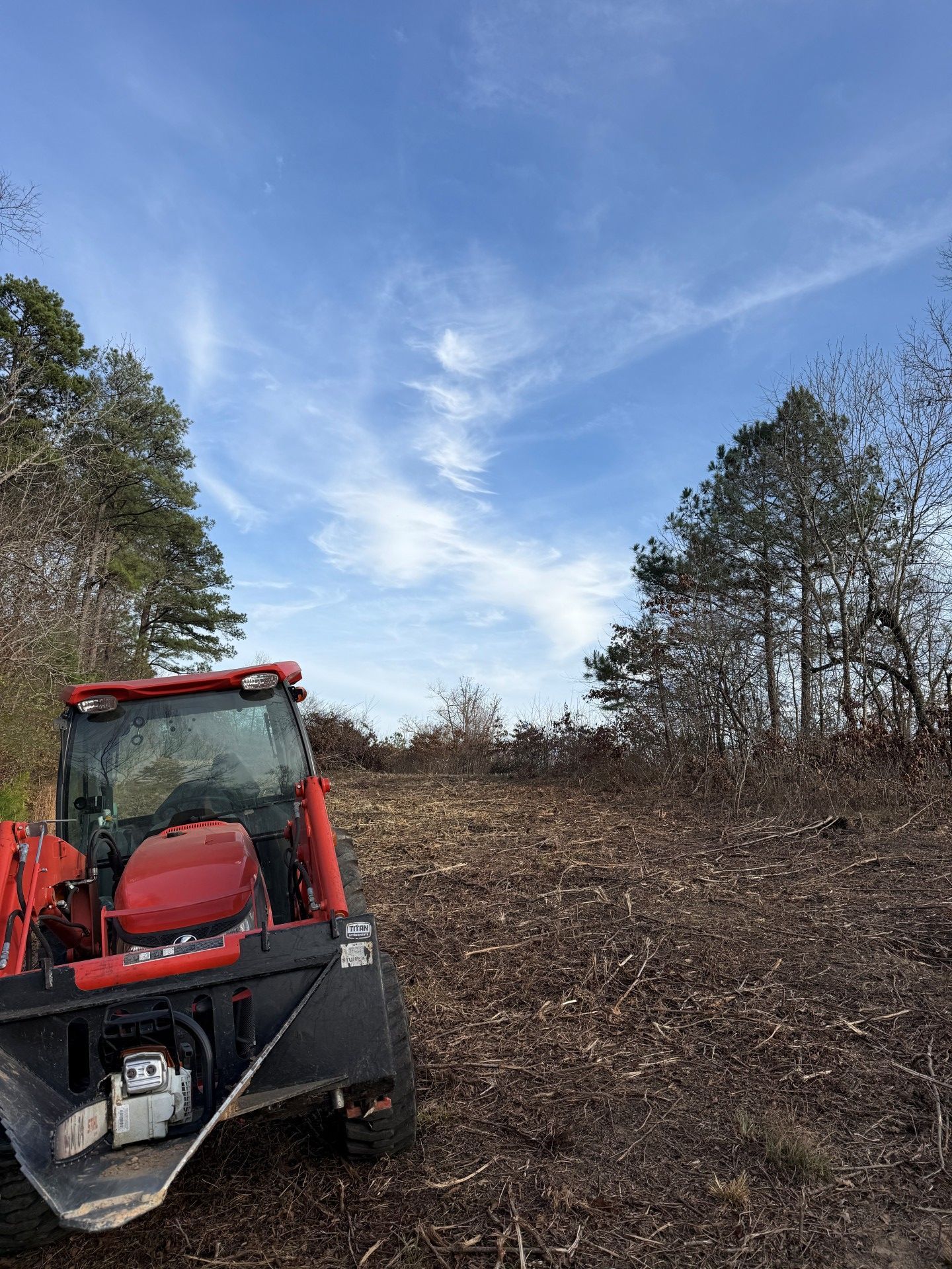 Red tractor clearing a wooded area under a blue sky with streaky clouds.