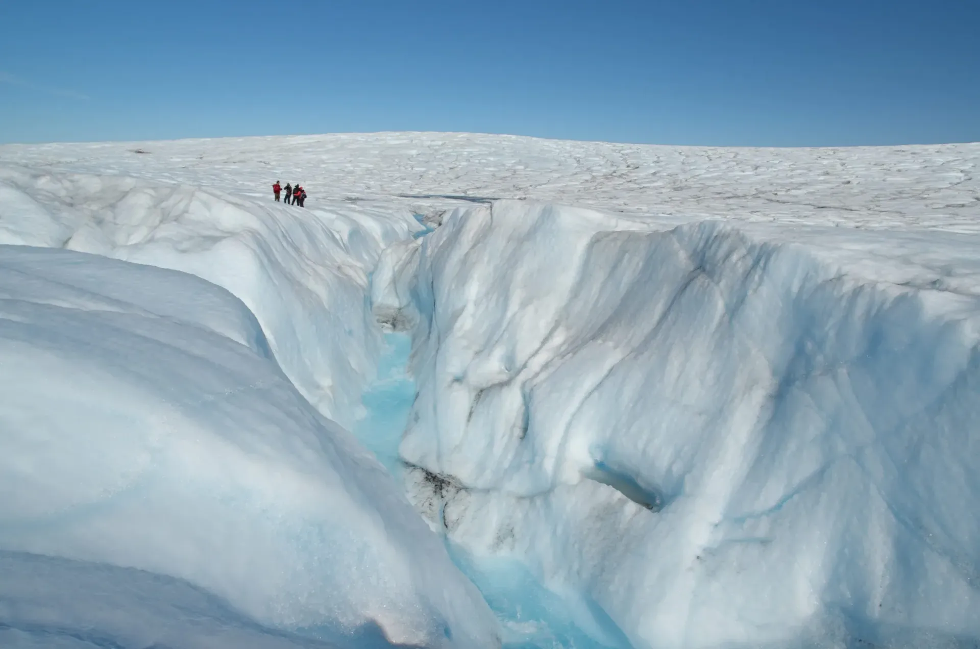 Mennesker står nær en dyb gletscherspalte; iskoldt blåt vand synligt i sprækken.