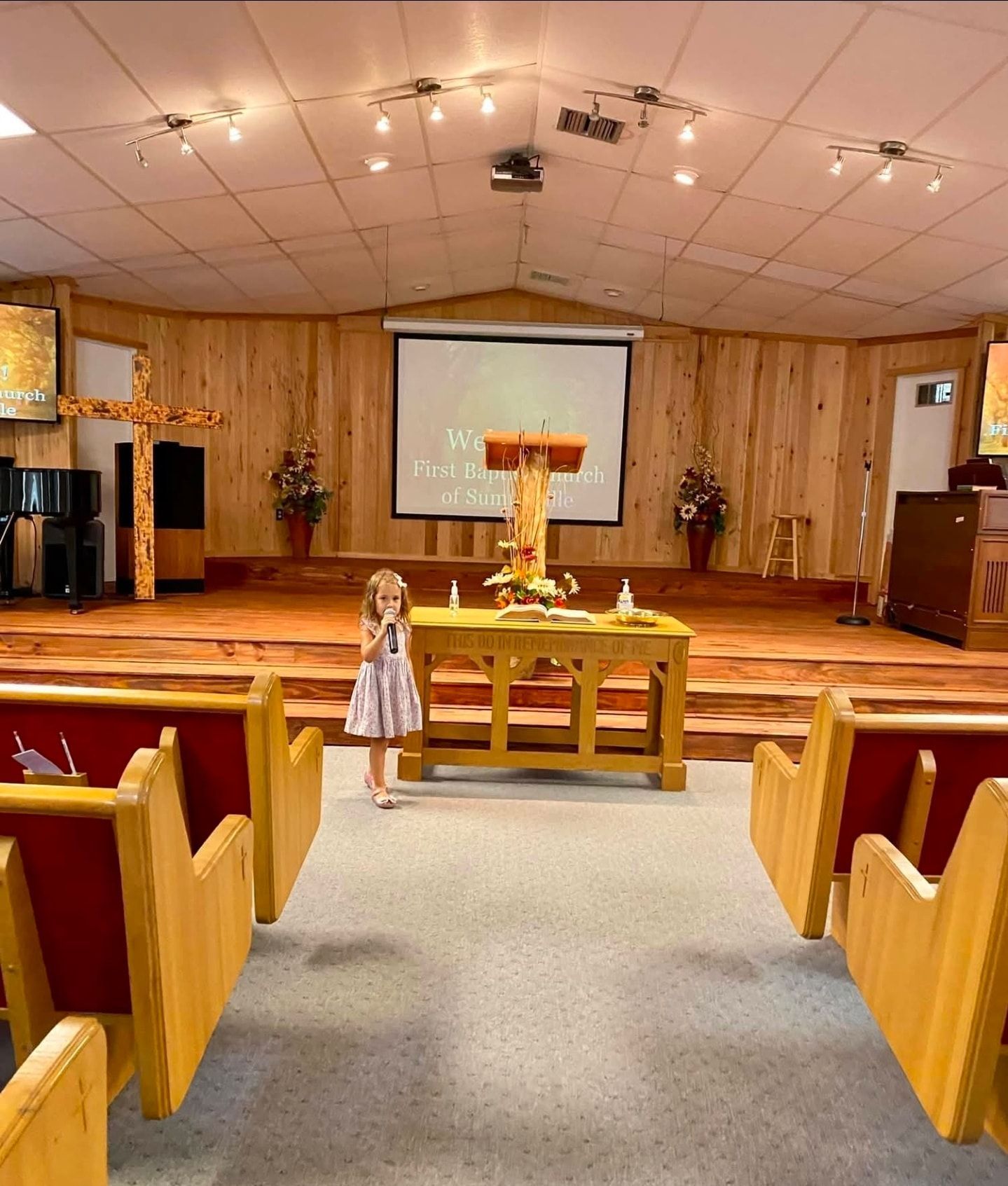 Child stands in church. Wooden altar, pews, stage with cross and screen visible.