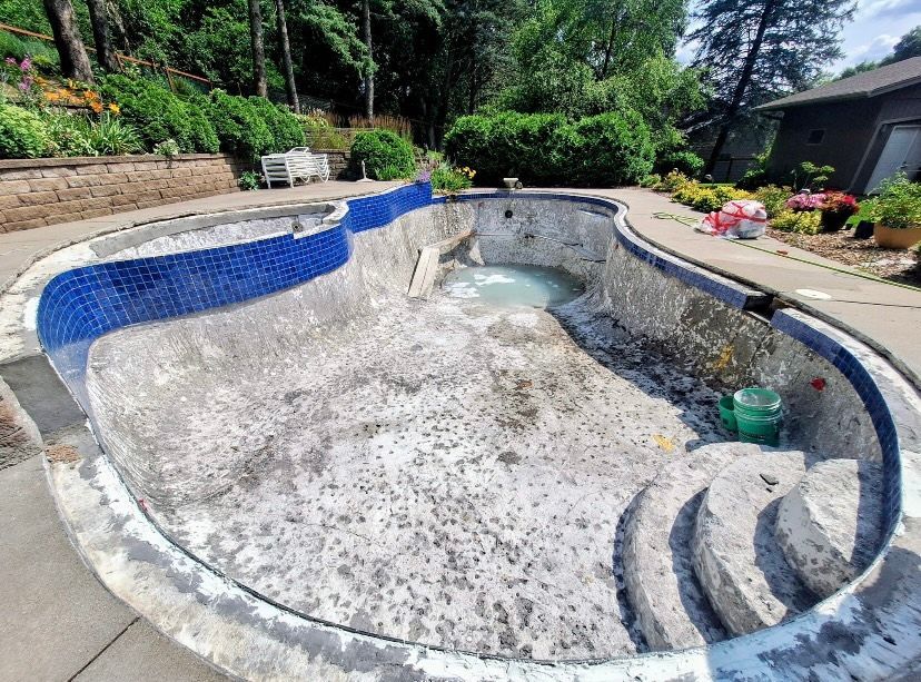 An empty backyard swimming pool undergoing renovation, showing exposed concrete, blue tile borders, and a set of stairs.