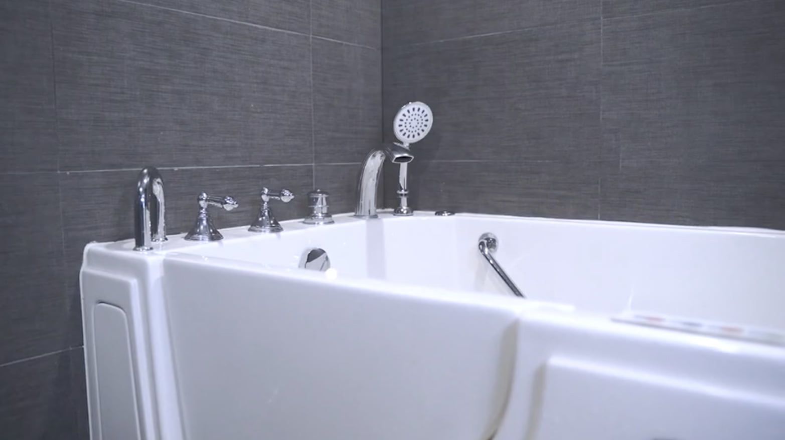 White walk-in bathtub with silver fixtures in a bathroom with gray tile walls.
