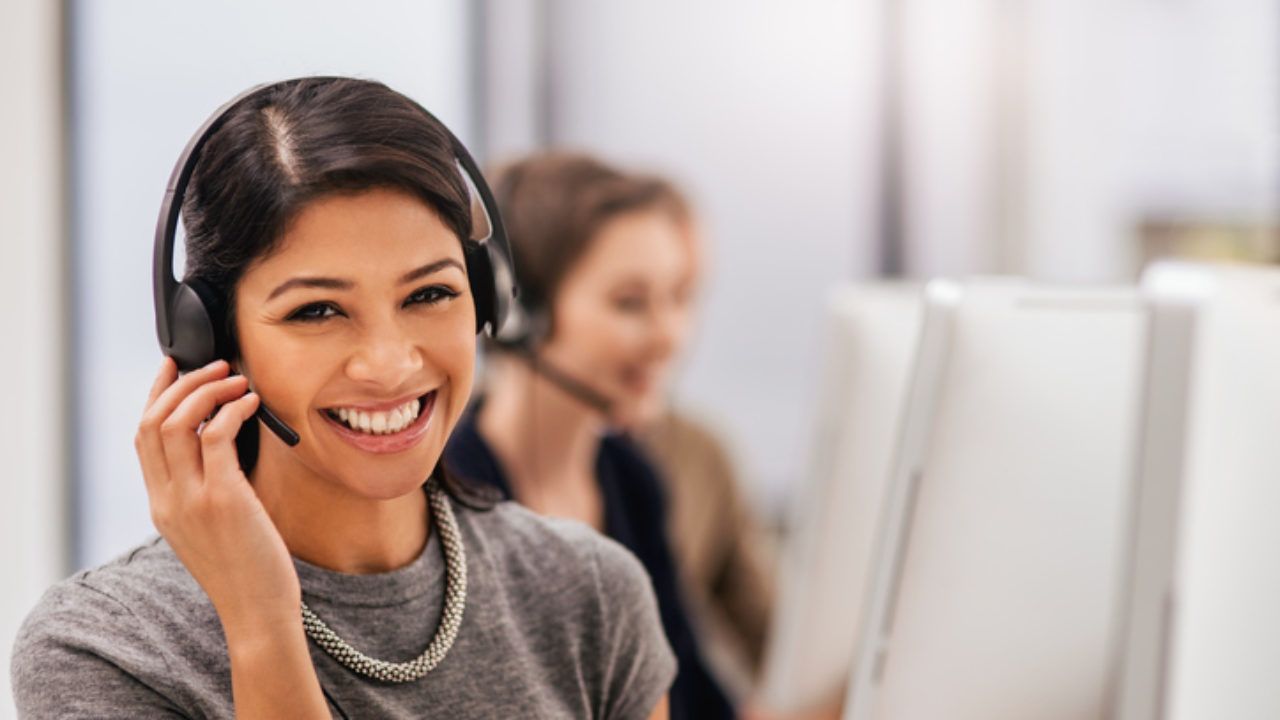 A woman wearing a headset is smiling while sitting in front of a computer.