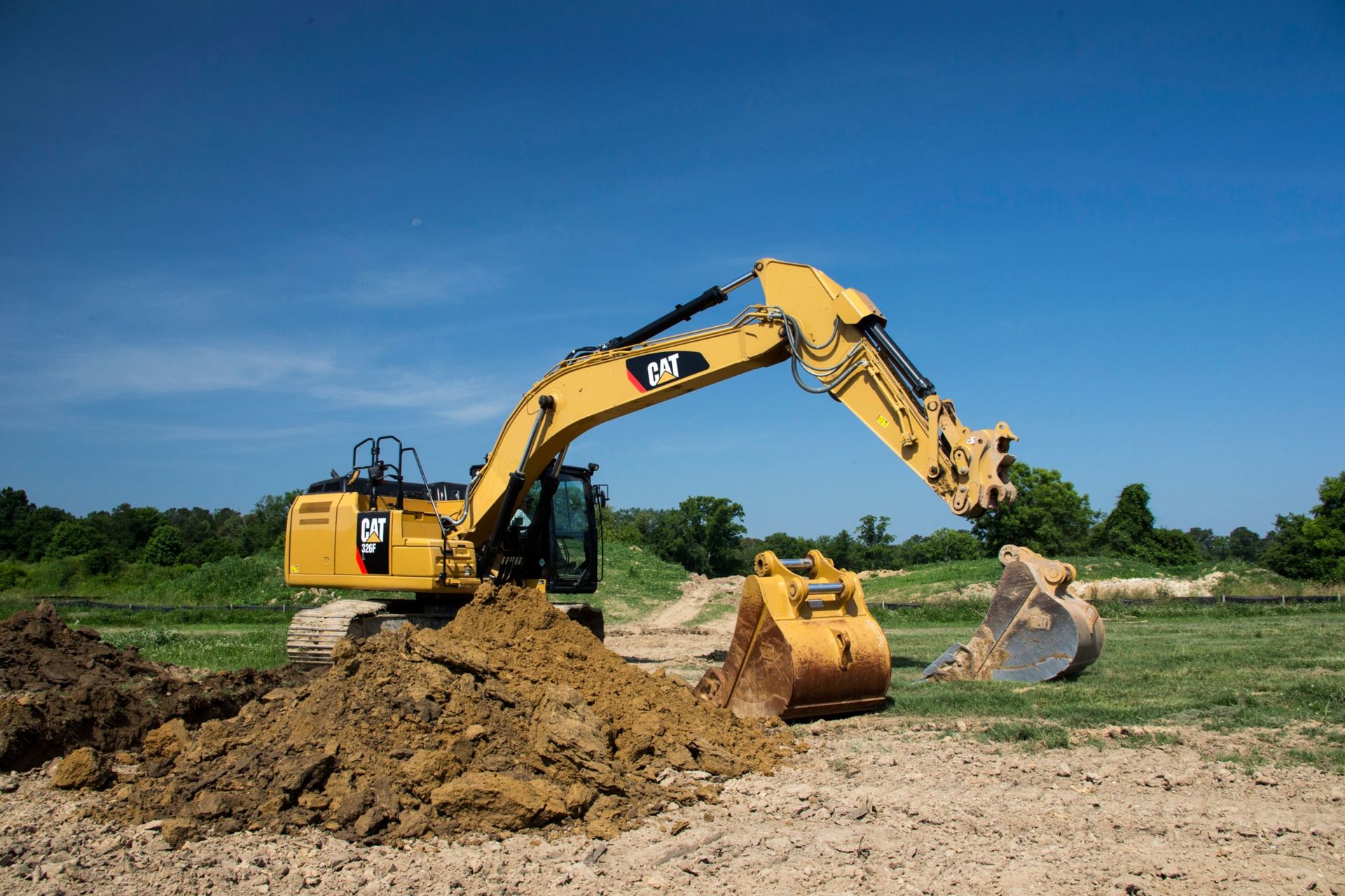 A DEV Builders yellow Caterpillar excavator digs into the earth under a clear blue sky. The machine is actively moving dirt.