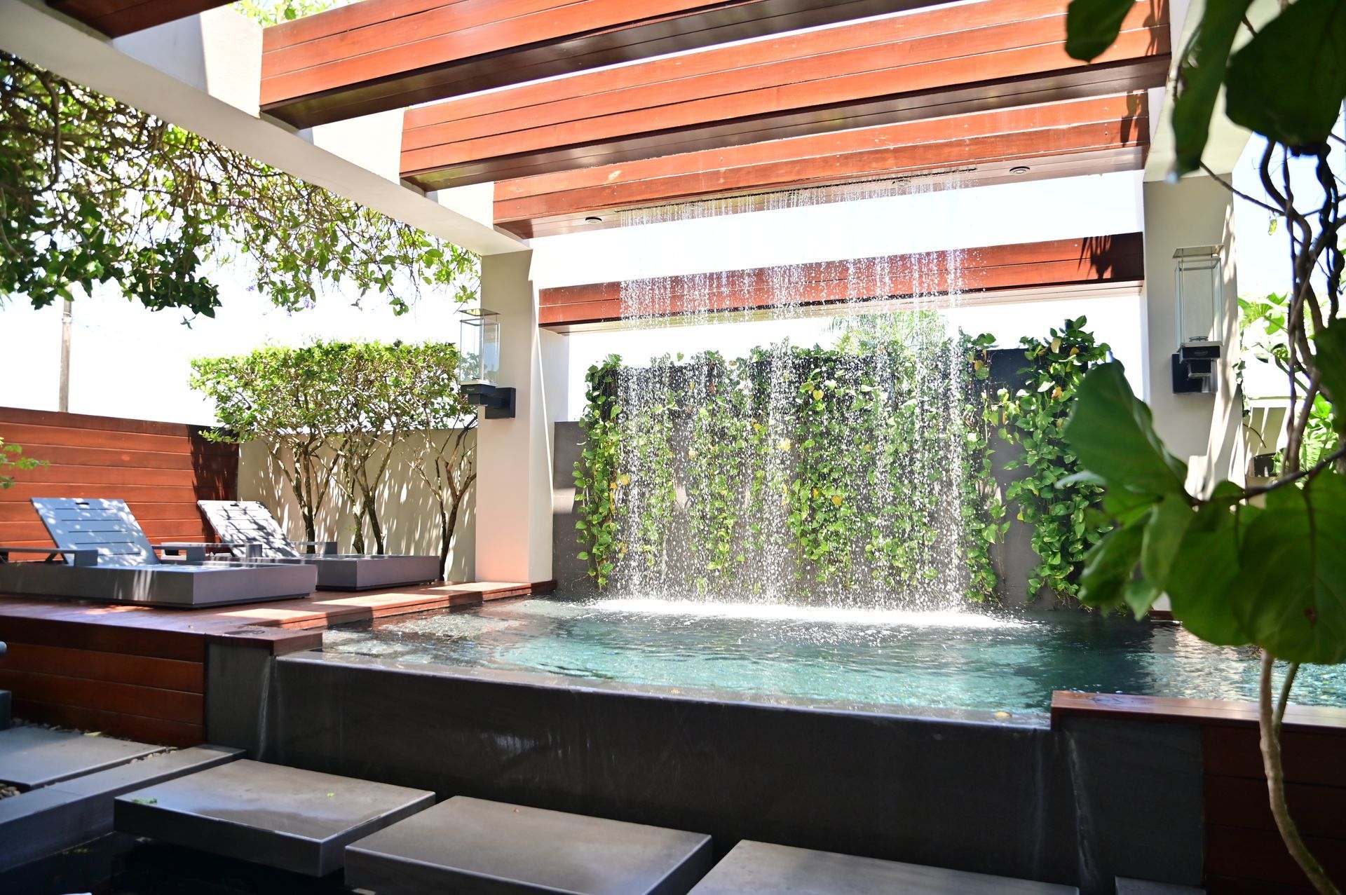 Pool with a waterfall feature surrounded by green foliage, under a wooden pergola.