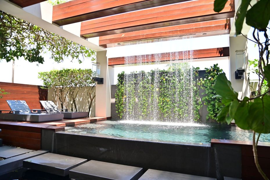 Pool with waterfall feature and wooden pergola, surrounded by greenery and seating.