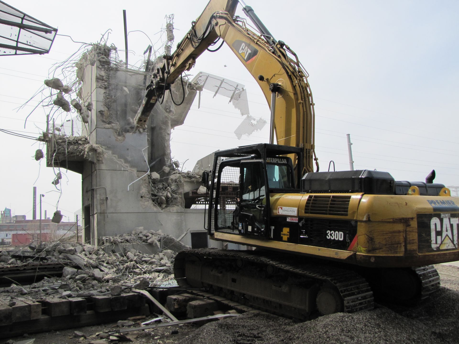 A DEV Builders worker in a yellow Caterpillar excavator demolishes a concrete structure, with debris scattered around.