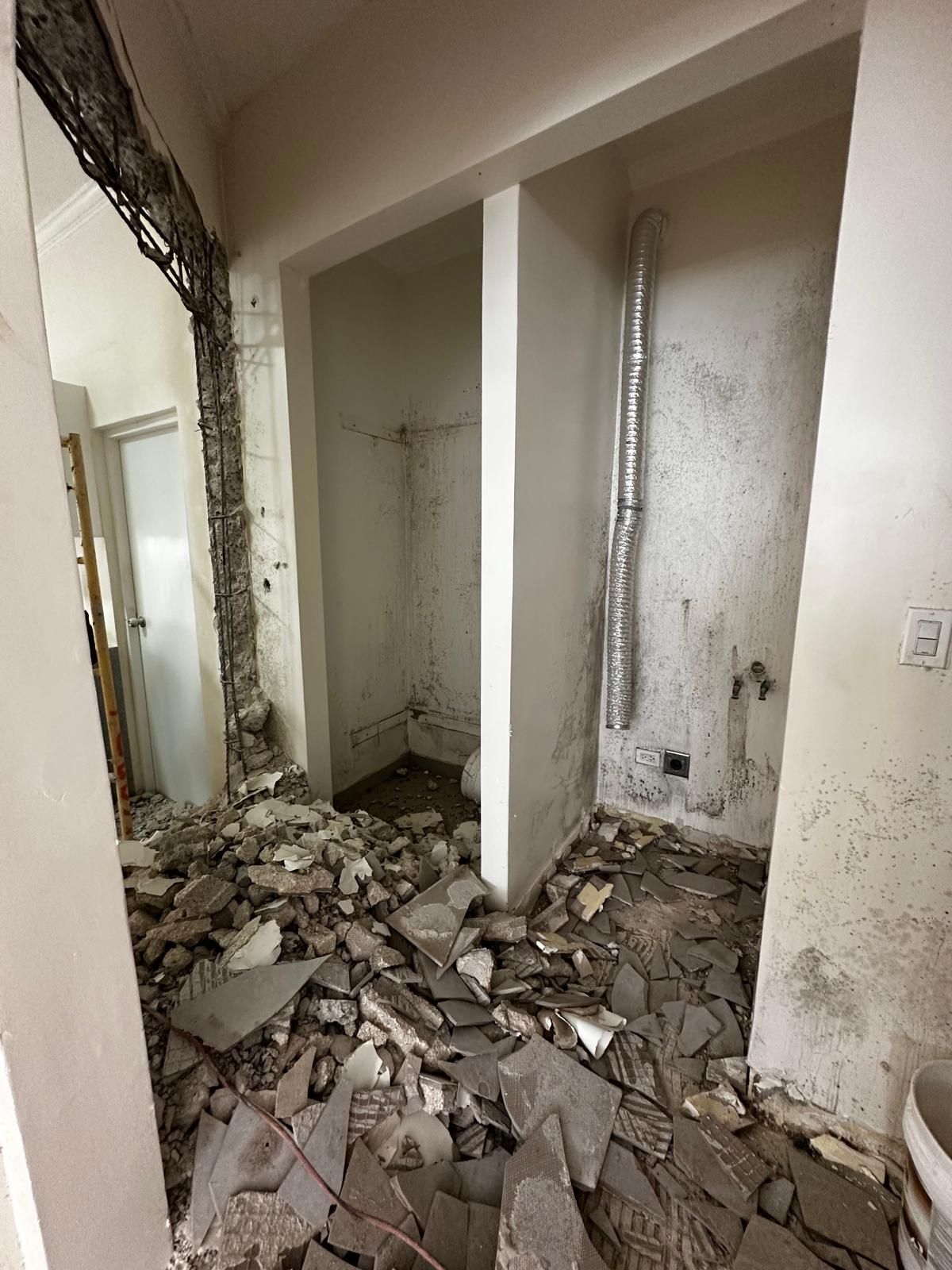 Demolished interior of a room. Debris litters the floor; an alcove with a toilet and a ventilation pipe is visible. Demolition done by DEV Builders Group.