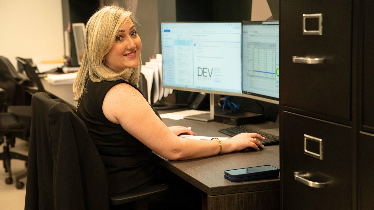A DEV Builders' worker is sitting at a desk in front of two computer monitors at DEV Builders Headquarters.