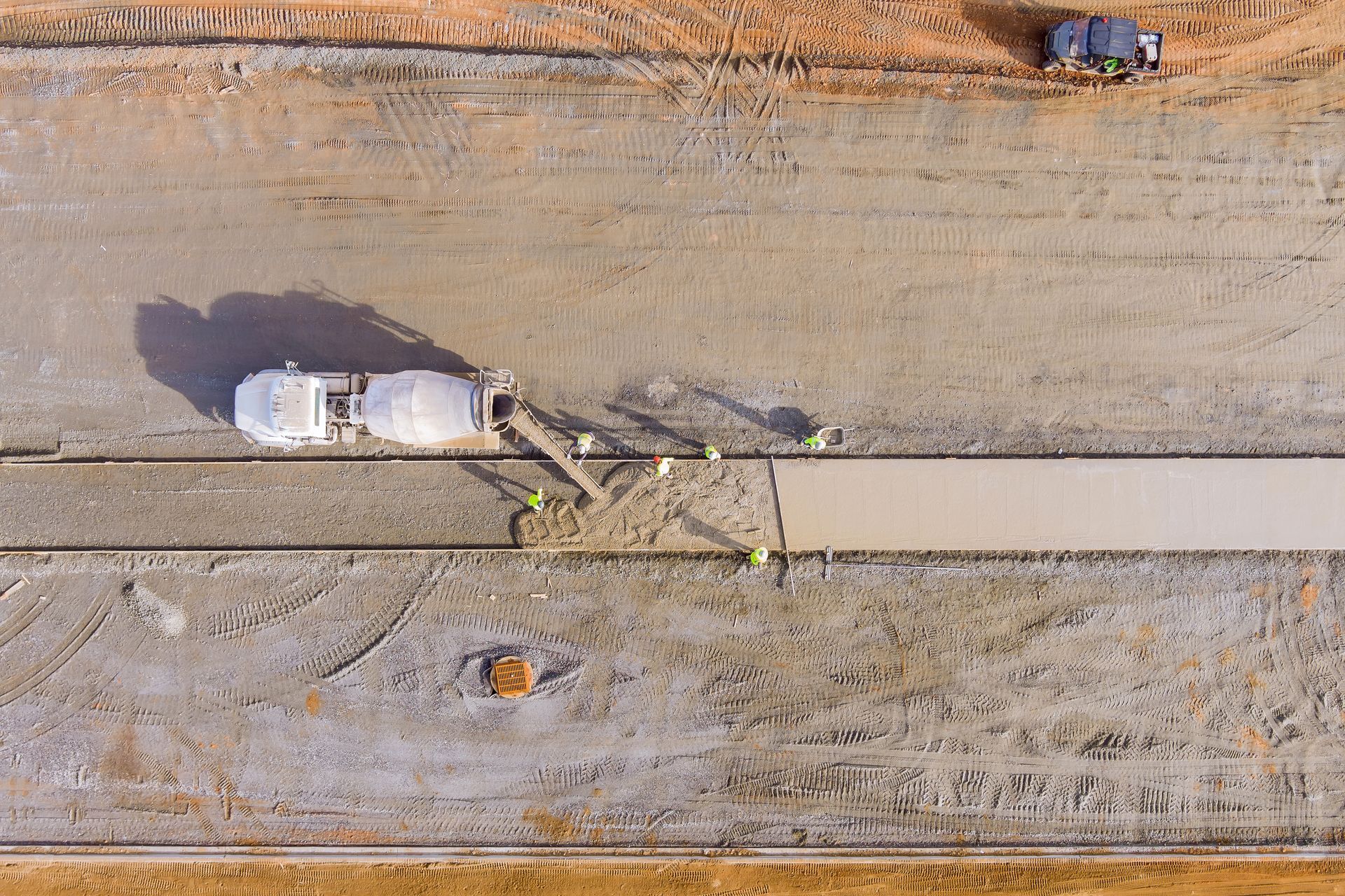 Aerial view of road construction: A cement mixer pours concrete onto a prepared strip as DEV Builders workers in yellow vests guide the process.