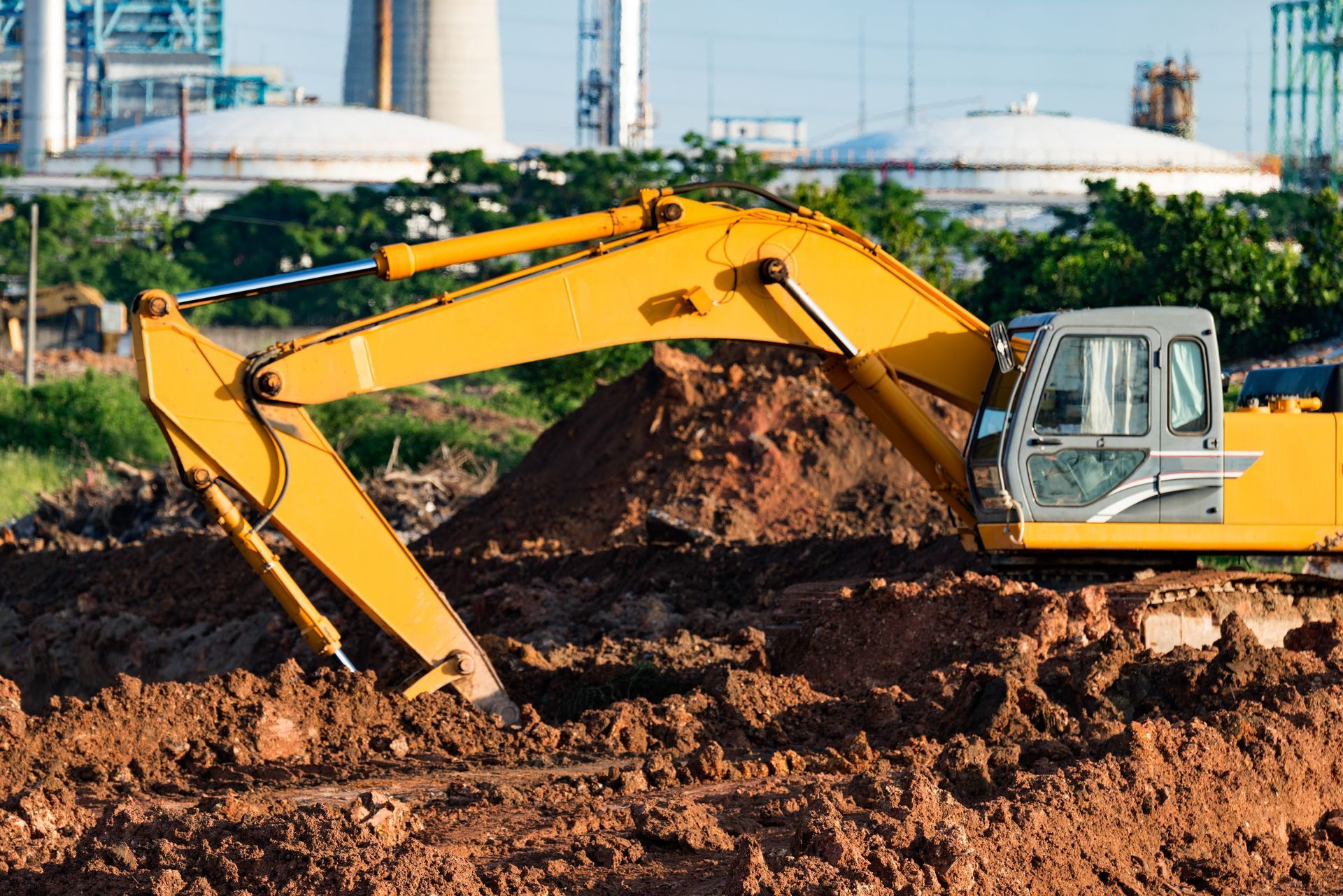 A DEV Builders yellow excavator digging in a brown dirt pile at a construction site with industrial buildings in the background.