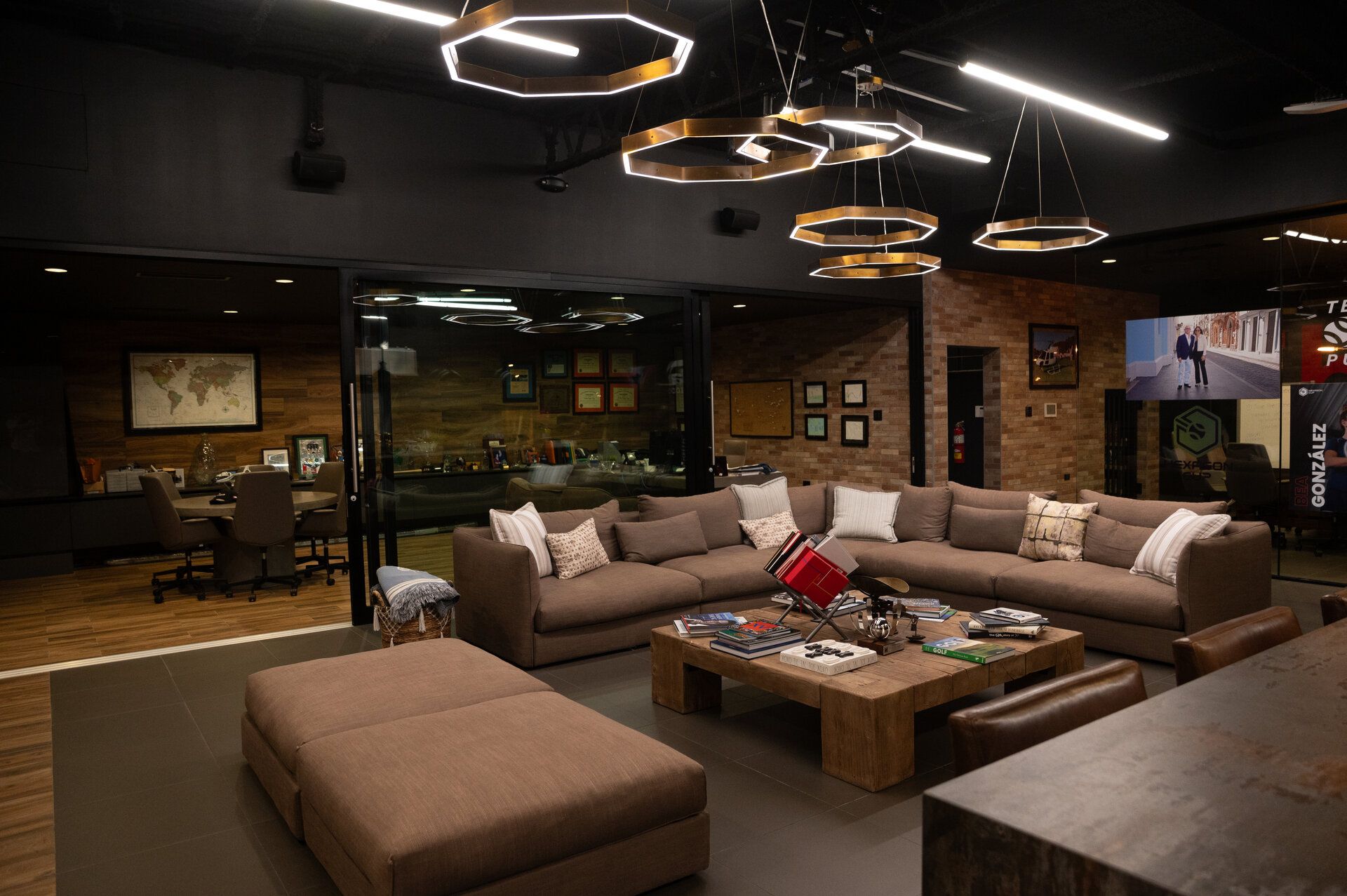 Modern lounge area with brown sectional sofa, ottoman, wooden coffee table, brick wall, and open office behind glass.