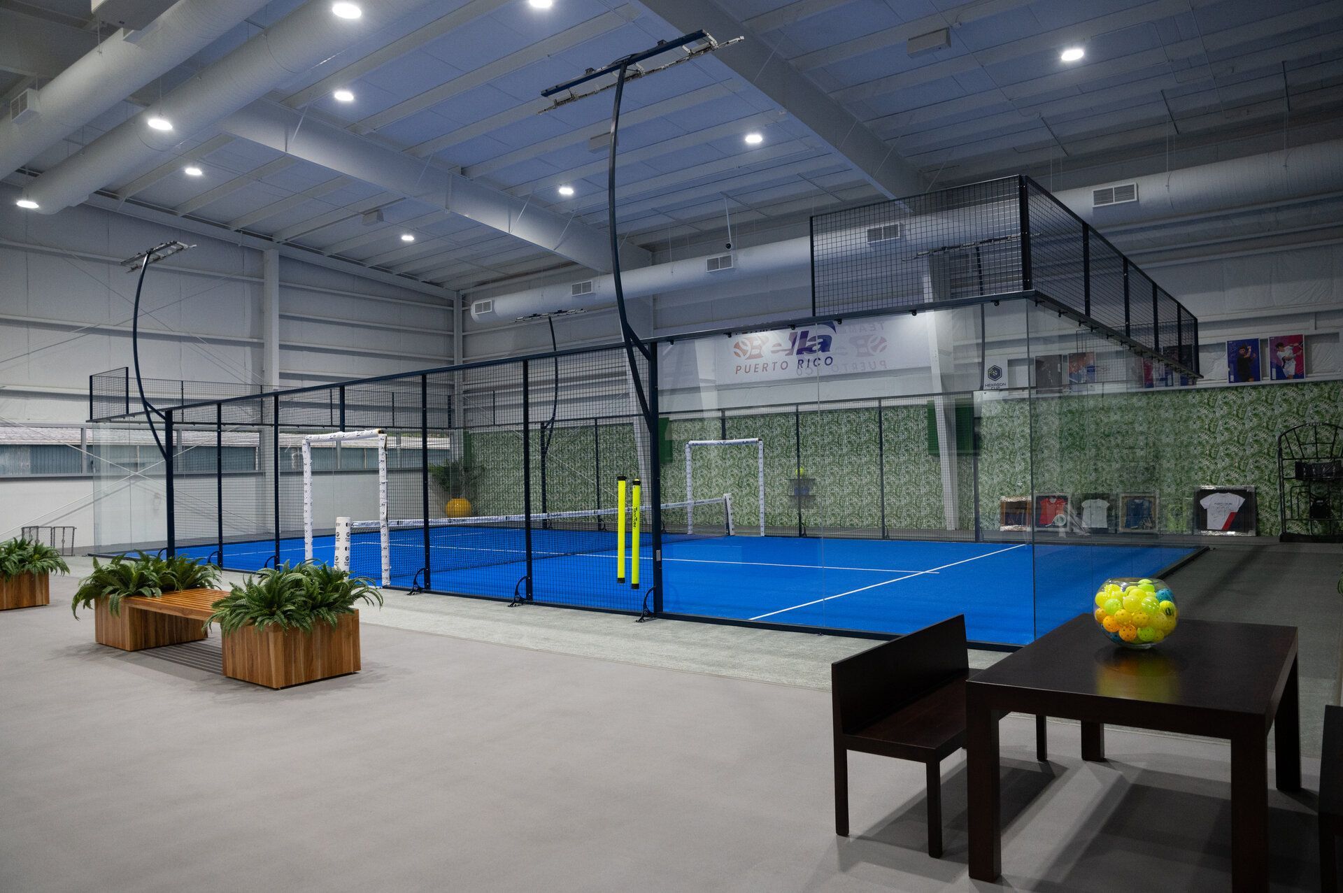 Indoor pickleball court in Puerto Rico with blue surface, surrounded by glass walls and netting.  Tables and benches are in the foreground.