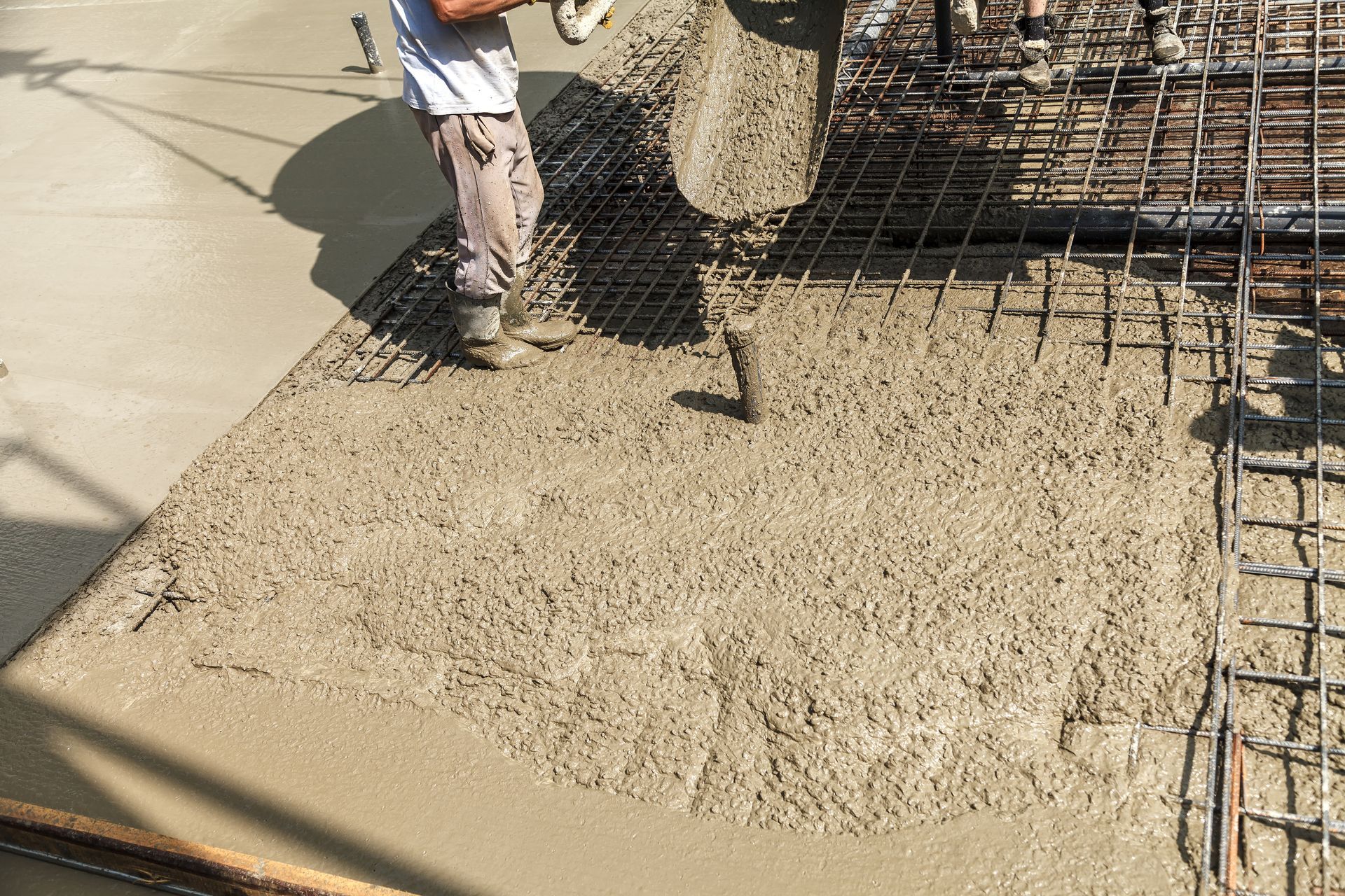 A DEV Builder construction worker pours wet concrete onto rebar framework, partially covering it. Sunny outdoor setting.