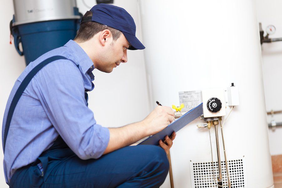 A man is kneeling down next to a water heater and writing on a clipboard.