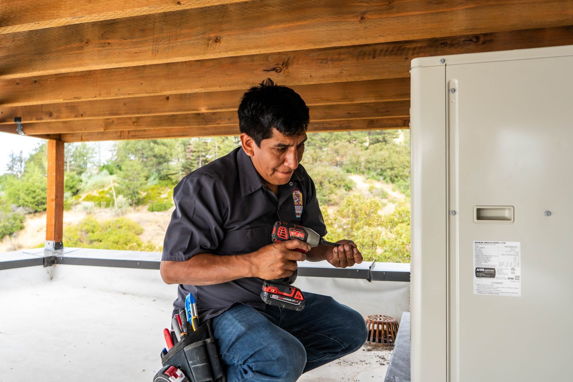 Lucas Heating & Air HVAC technician kneels, using a drill on an outdoor unit, on a rooftop.