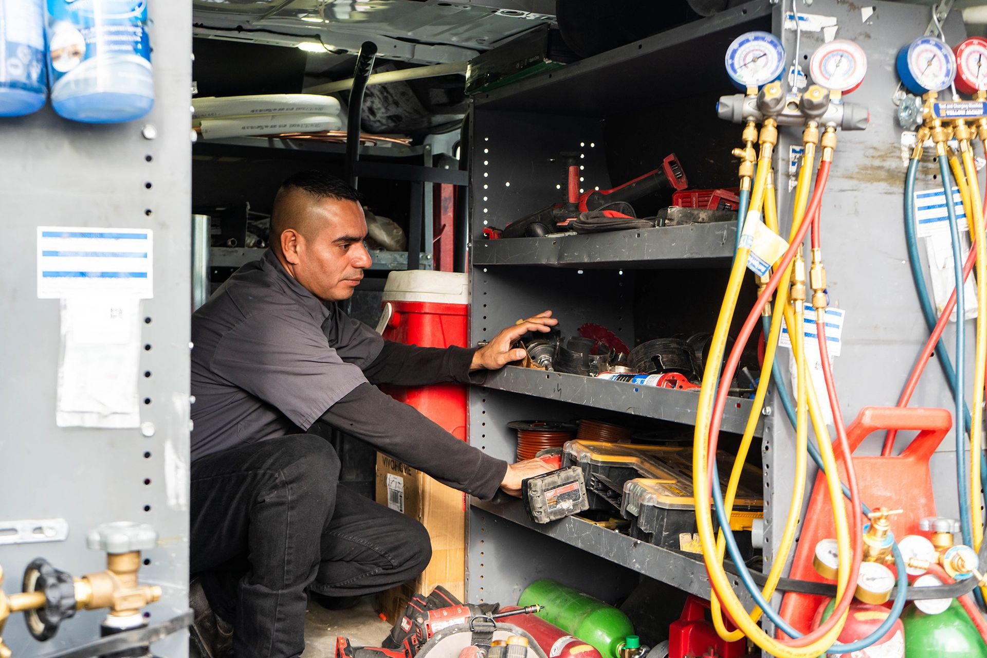 Lucas Heating & Air HVAC technician in a van, crouched, reaching for tools on shelves. Gauges, tanks, and equipment visible.