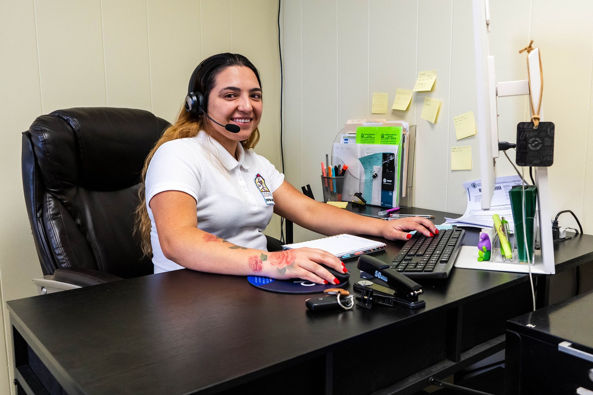 Woman with headset at a desk, smiling. Typing on keyboard, with stapler and keys. Office setting.