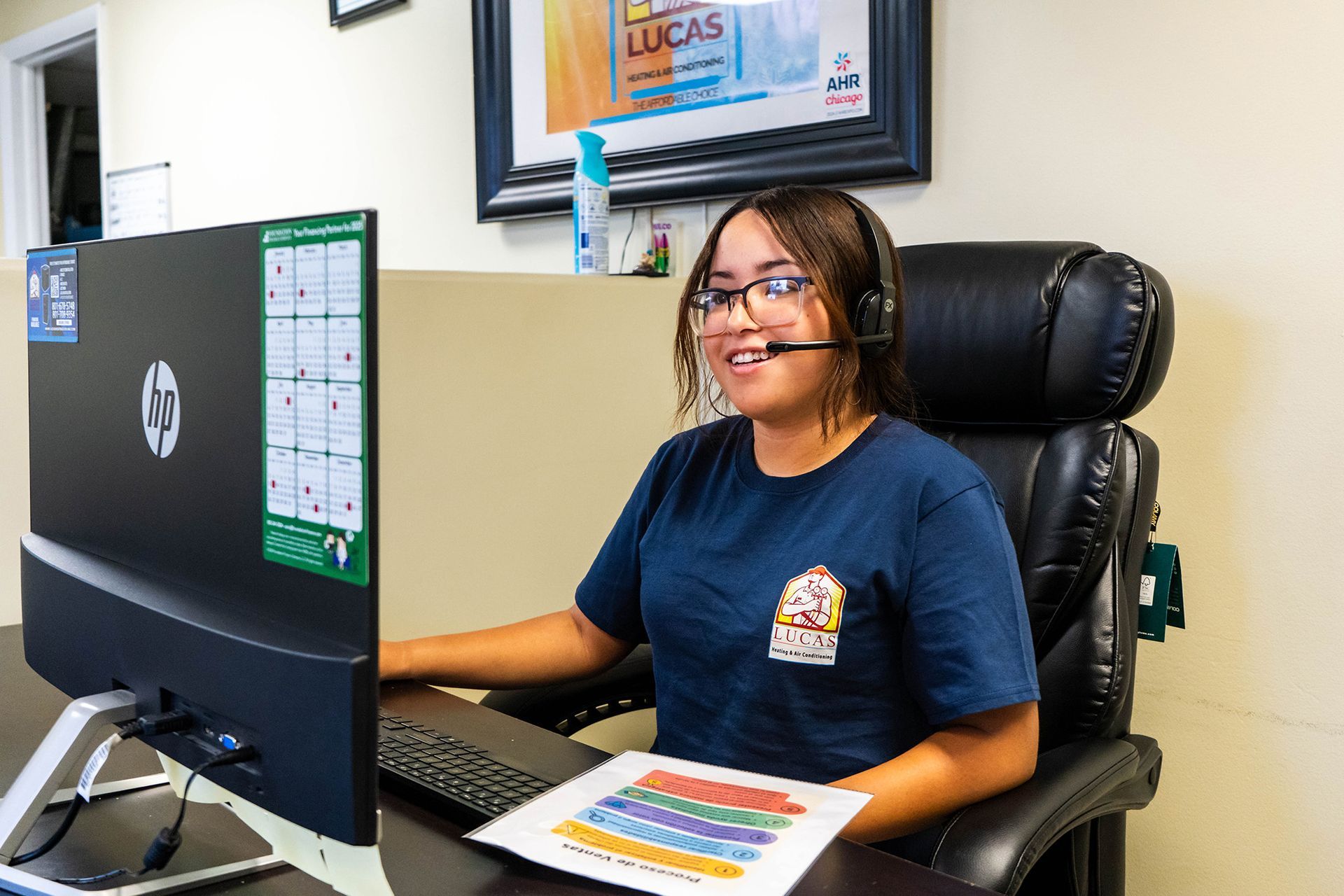 A Lucas Heating & Air employee wearing a headset smiles while working at a computer in an office.