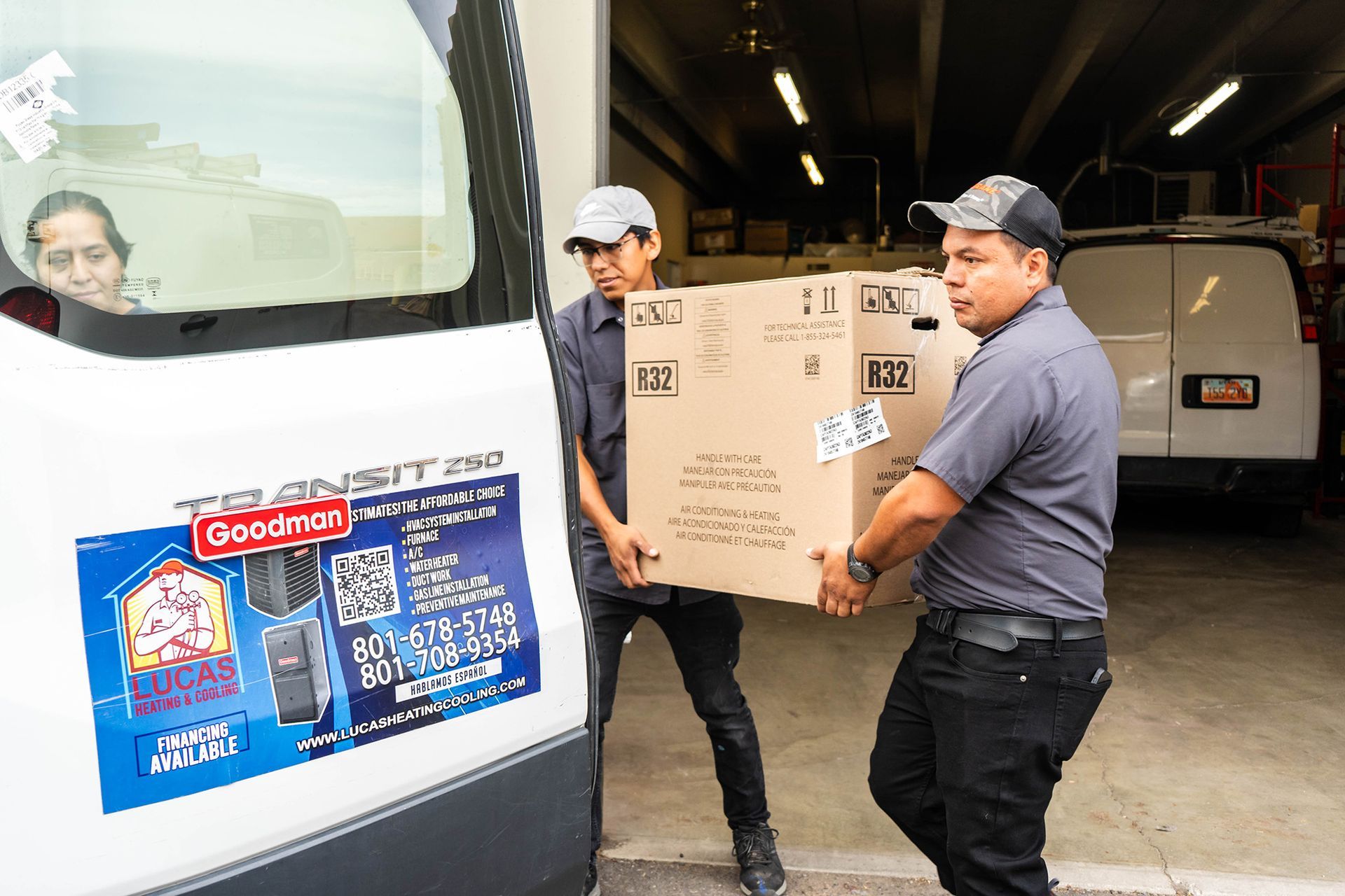 Two men carrying a large cardboard box from a Goodman van. Woman in van looks on.