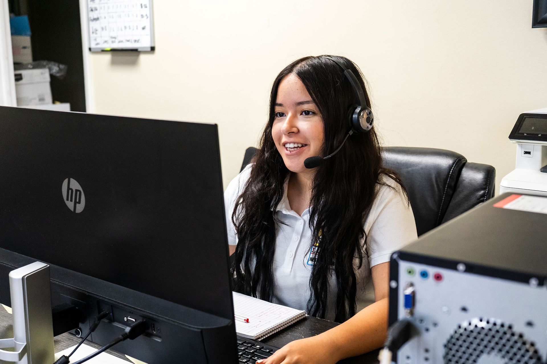 Woman with a headset smiles while working on a computer in an office setting.