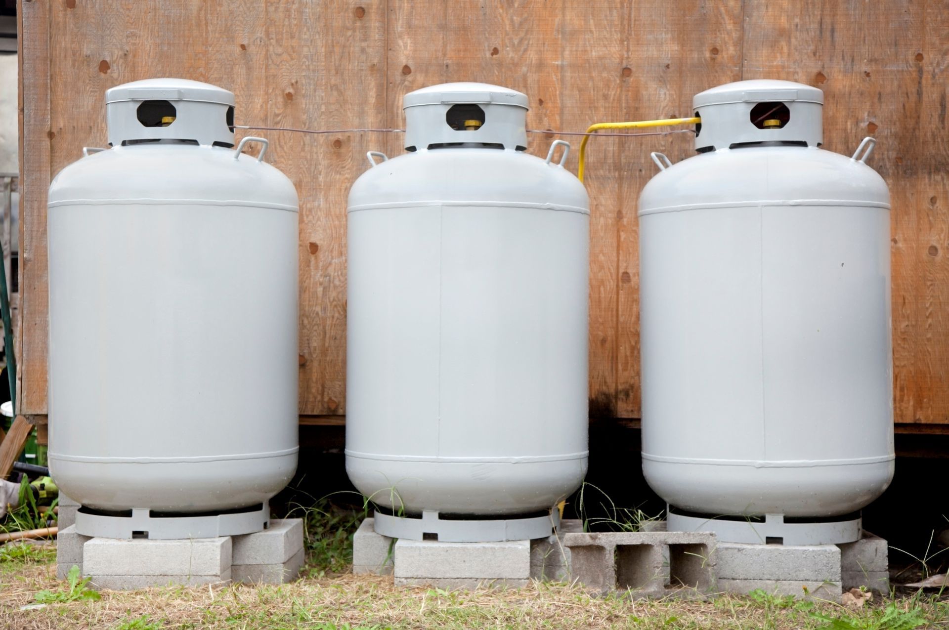 Three gray propane tanks on concrete blocks front of wood wall.
