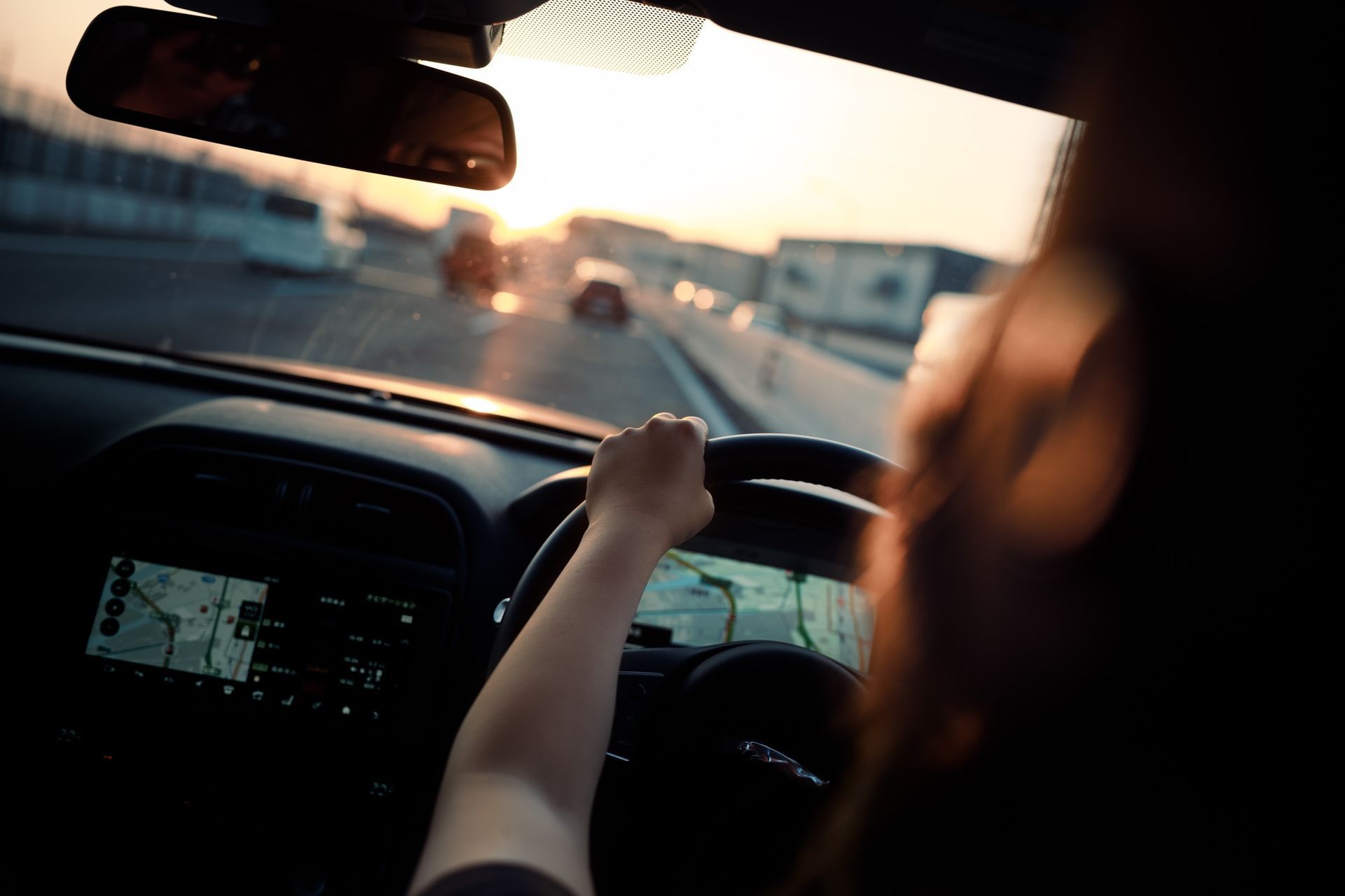 Person driving car on highway at sunset; hand on steering wheel, dashboard, and road visible.