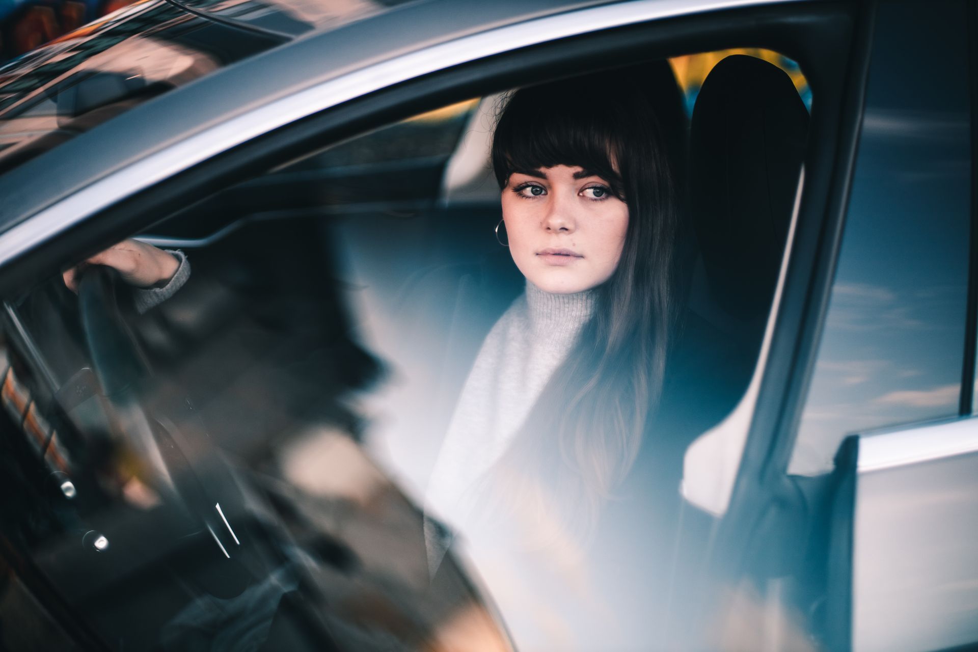Woman in car, looking out. Dark hair, bangs, neutral expression, gray car.