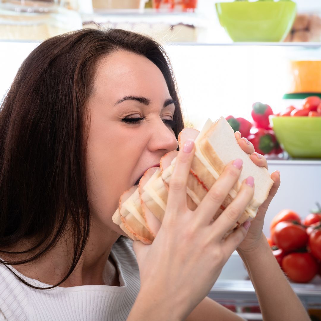 A woman is eating a sandwich in front of a refrigerator full of fruits and vegetables