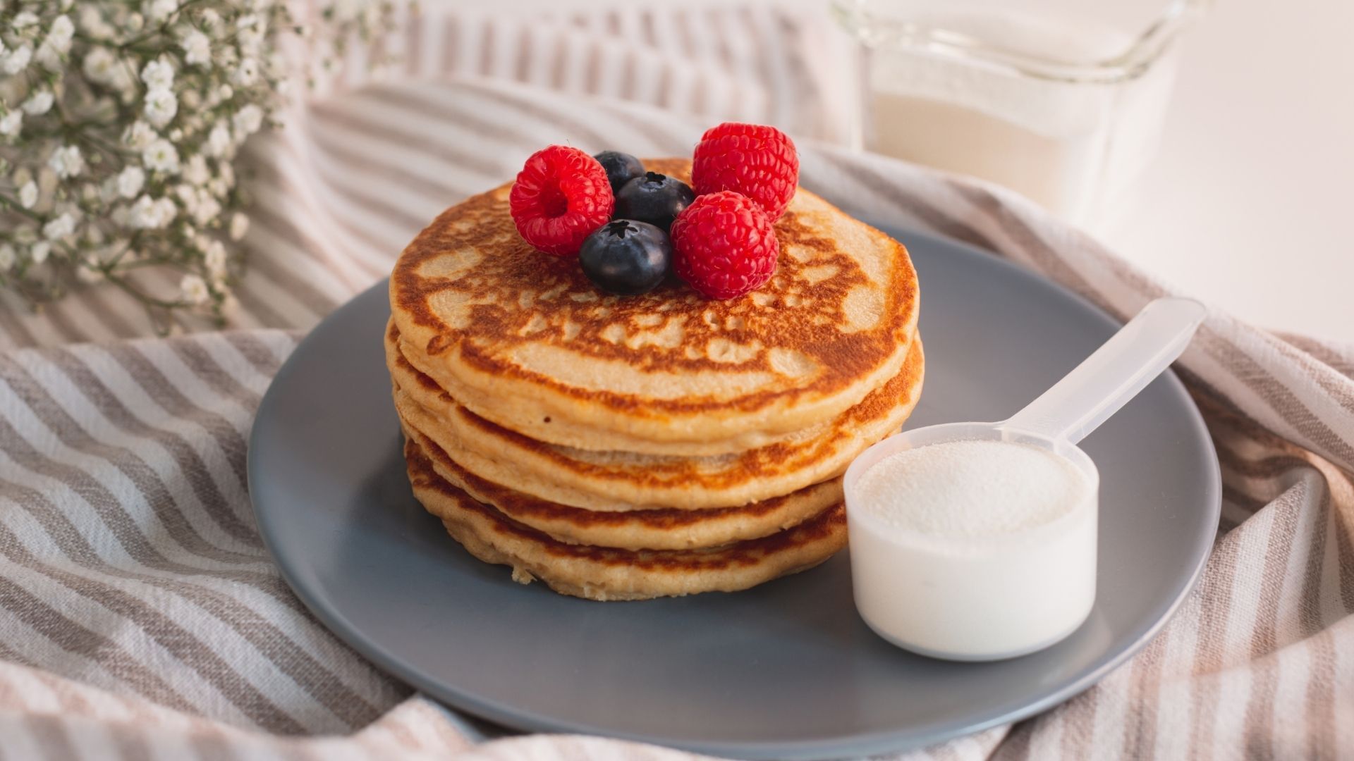 A stack of pancakes with raspberries and blueberries on a plate with a measuring cup of milk.