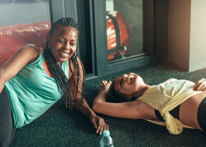 Two women are laying on the floor in a gym laughing.