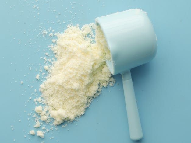 A scoop of milk powder is spilling out of it on a blue surface. Protein