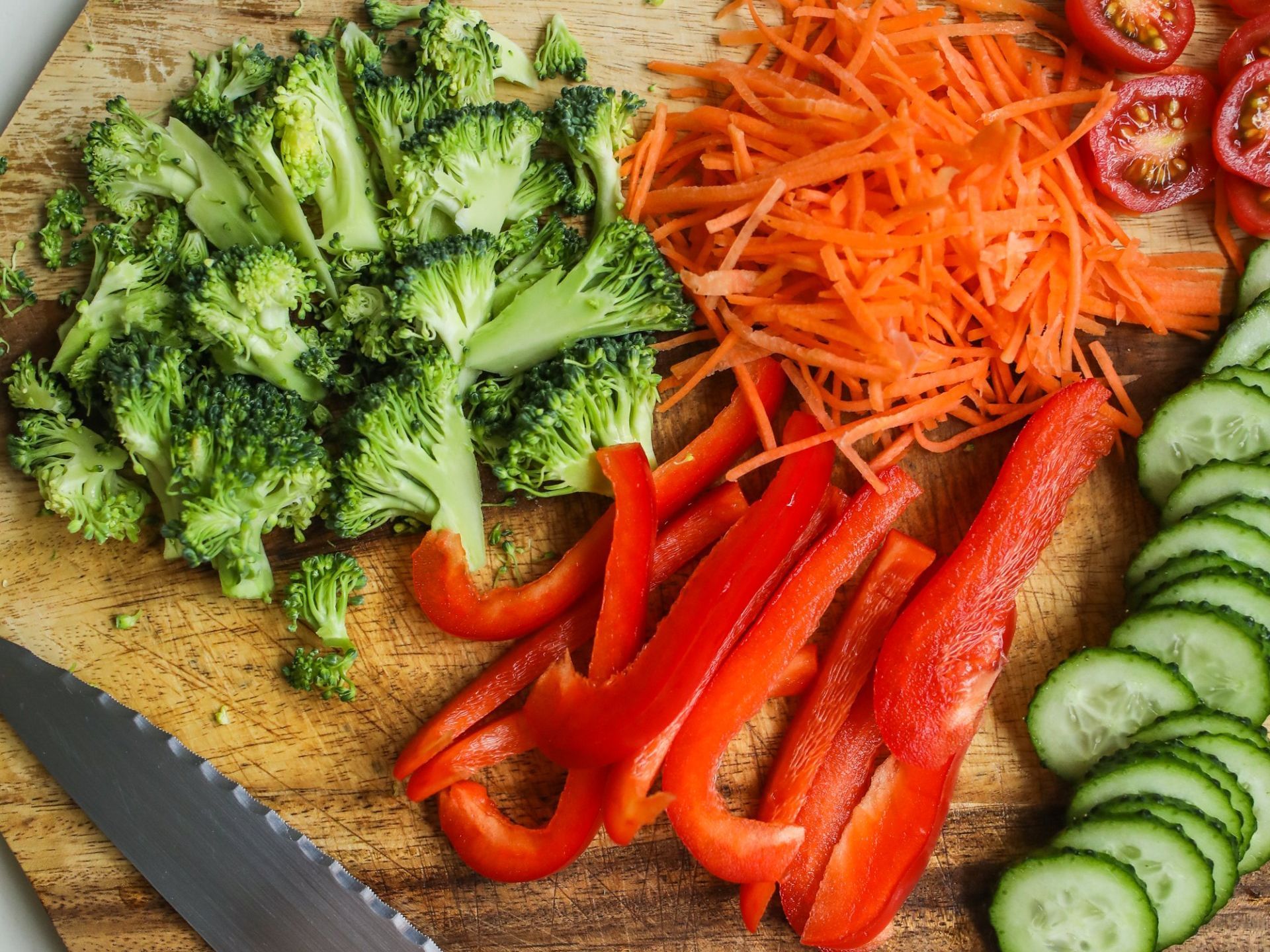 A wooden cutting board topped with broccoli , carrots , peppers , cucumbers and tomatoes.