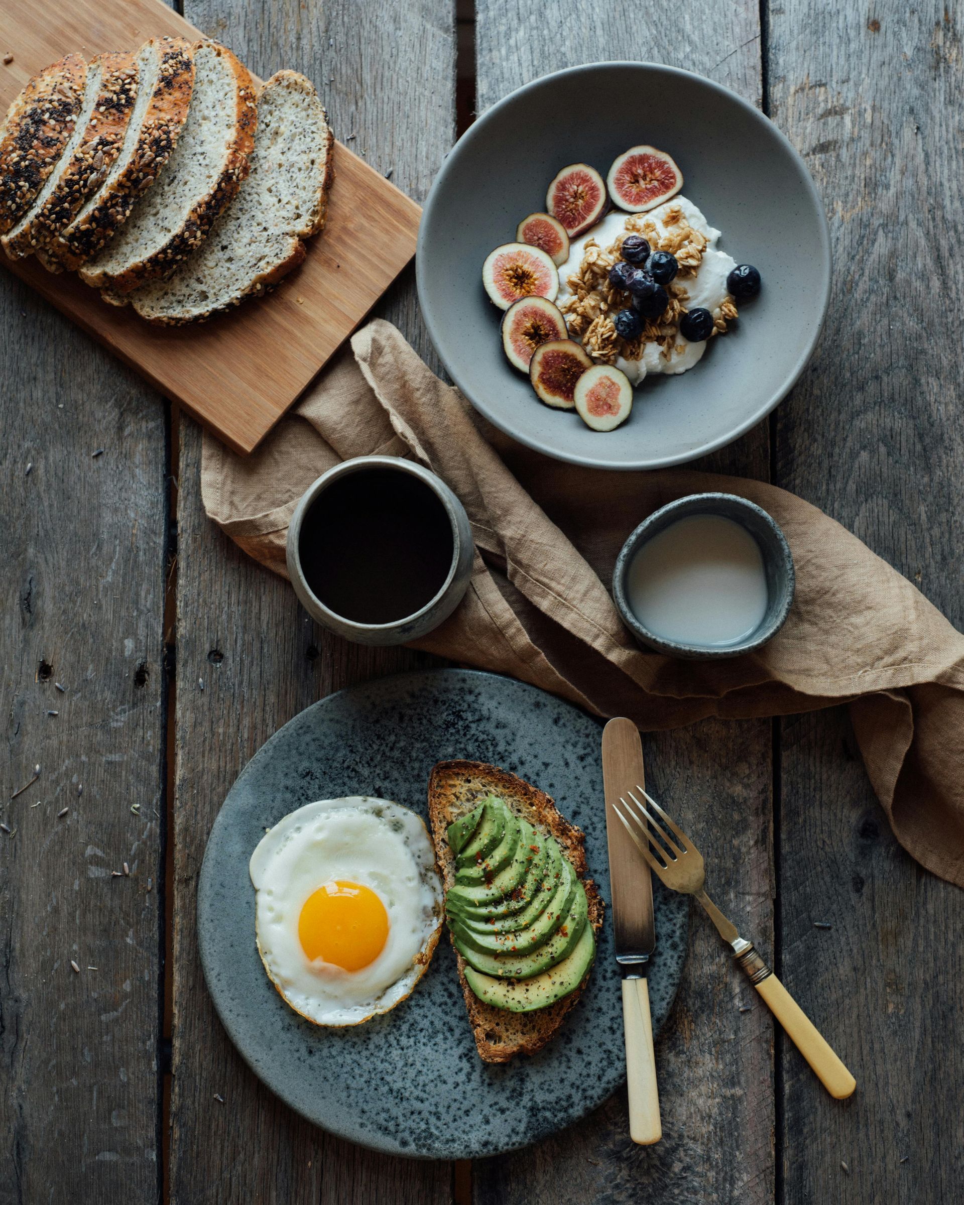 Breakfast spread: toast, eggs, avocado, yogurt with figs & blueberries, bread, coffee, and milk.
