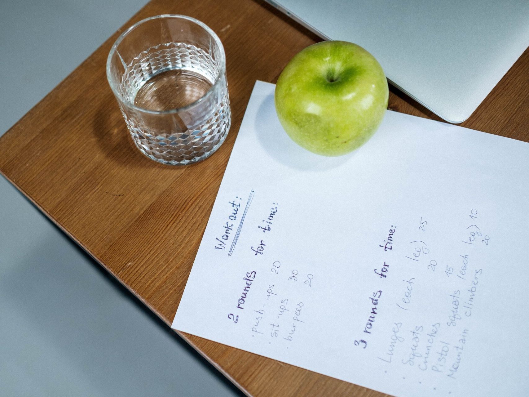 A green apple sits on a wooden table next to a glass of water