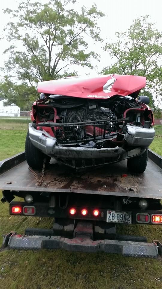 A severely damaged red car loaded onto a flatbed tow truck. The car's roof is caved in.
