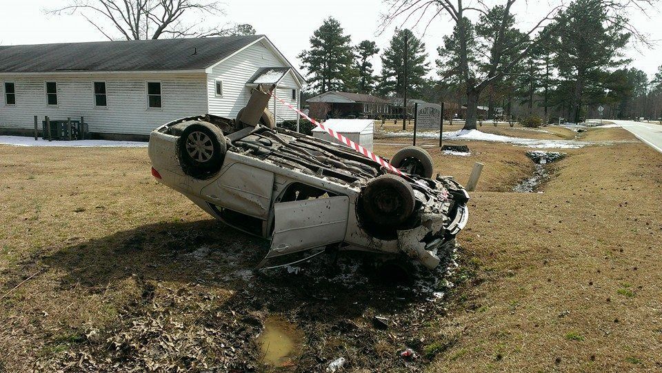 Car overturned in a ditch next to a road, near a building.