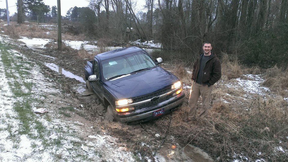 Truck stuck in muddy ditch next to a snowy field; man stands beside it.