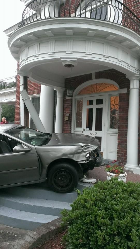 Car crashed into the entrance of a brick building with white columns. Gray car, damaged front, next to a white door.