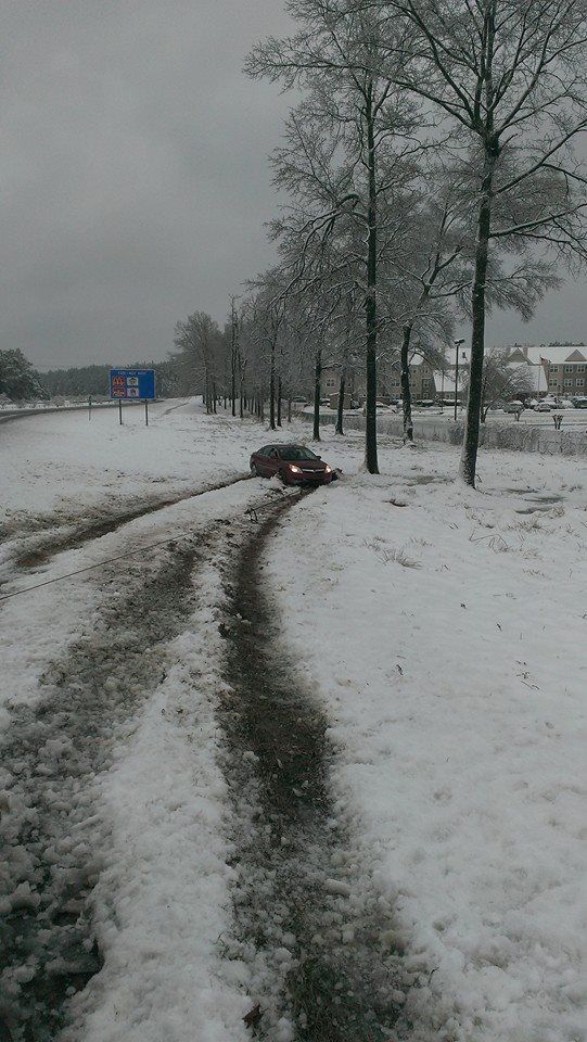 Car stuck in snow on the side of a road, next to trees. Overcast sky.