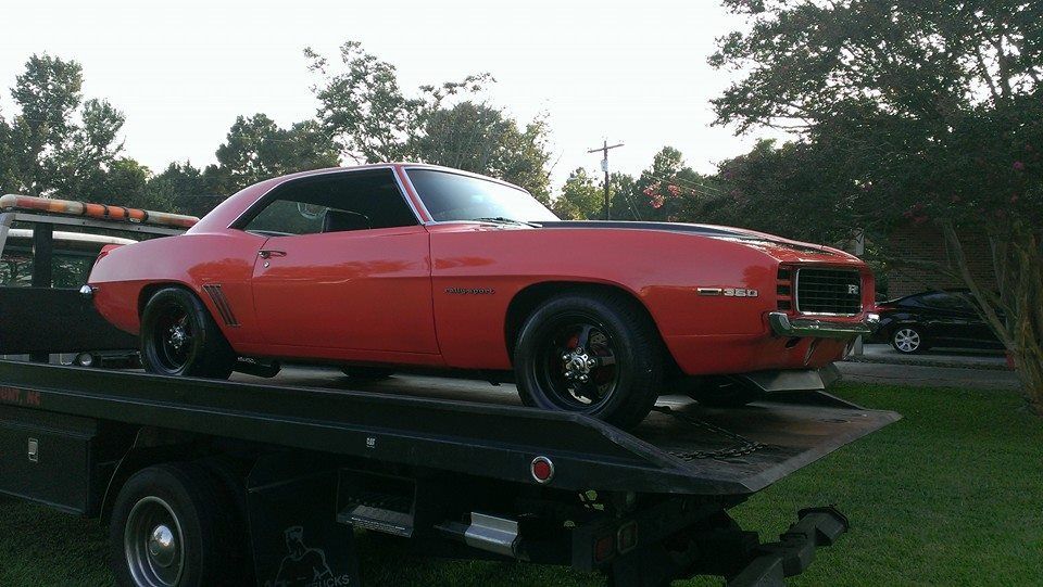 Red classic car on a tow truck, black wheels, dark hood stripes, parked outside during the day.