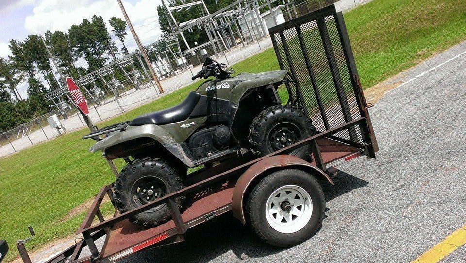 Green ATV on a trailer next to a paved road, in front of a power station.