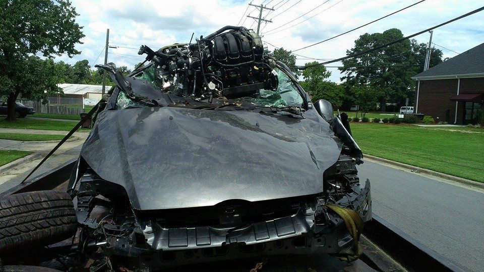 Severely damaged black car on a tow truck, with engine and roof destroyed, parked on a street.