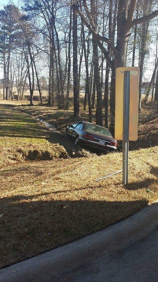 Car crashed into grassy ditch next to a yellow sign and trees.